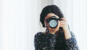 Woman in front of white background holding a camera to her face.