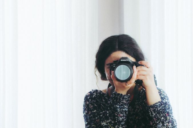 Woman in front of white background holding a camera to her face.